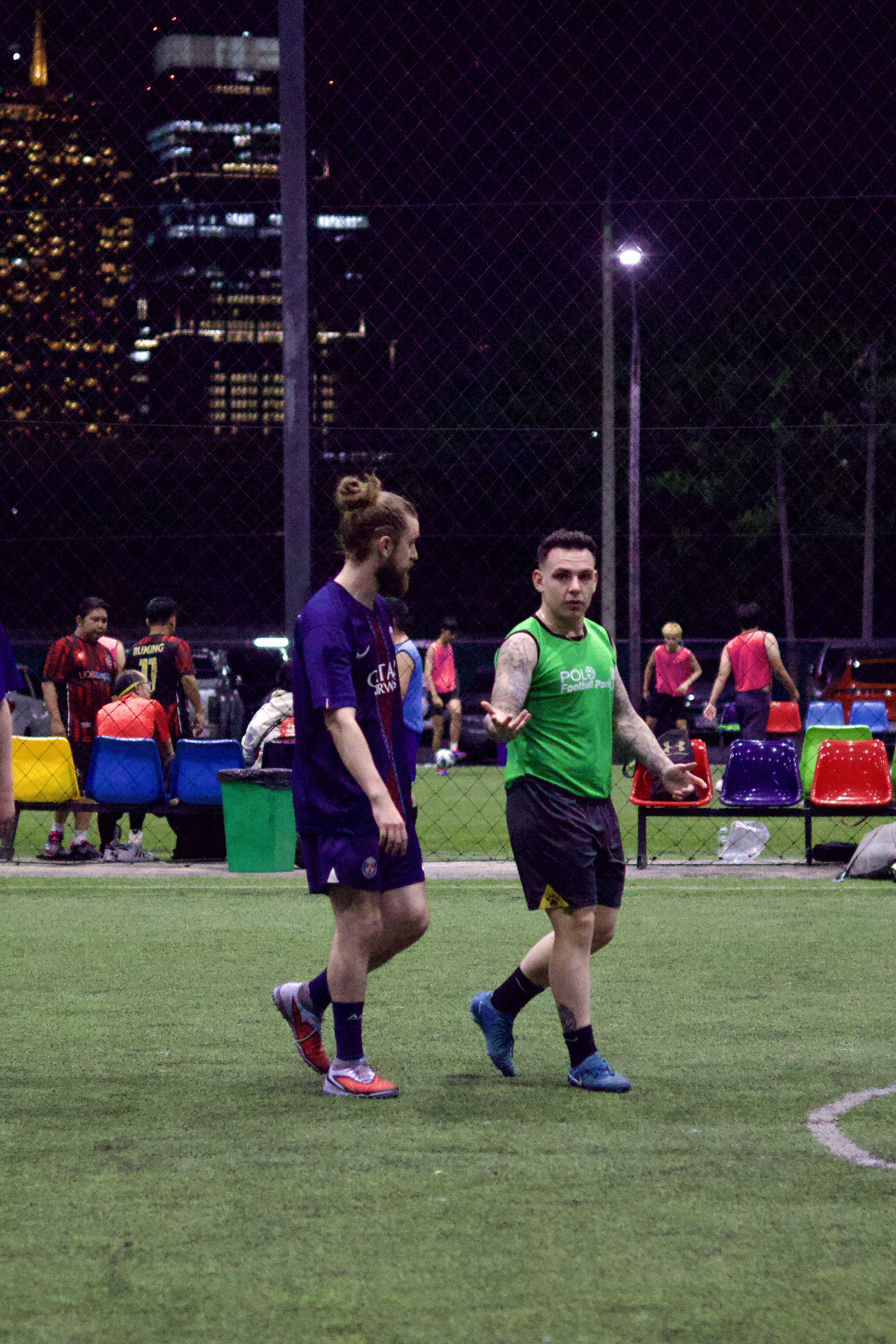 Football players on a synthetic turf pitch under evening lights during light rain in Bangkok with wet surface reflections