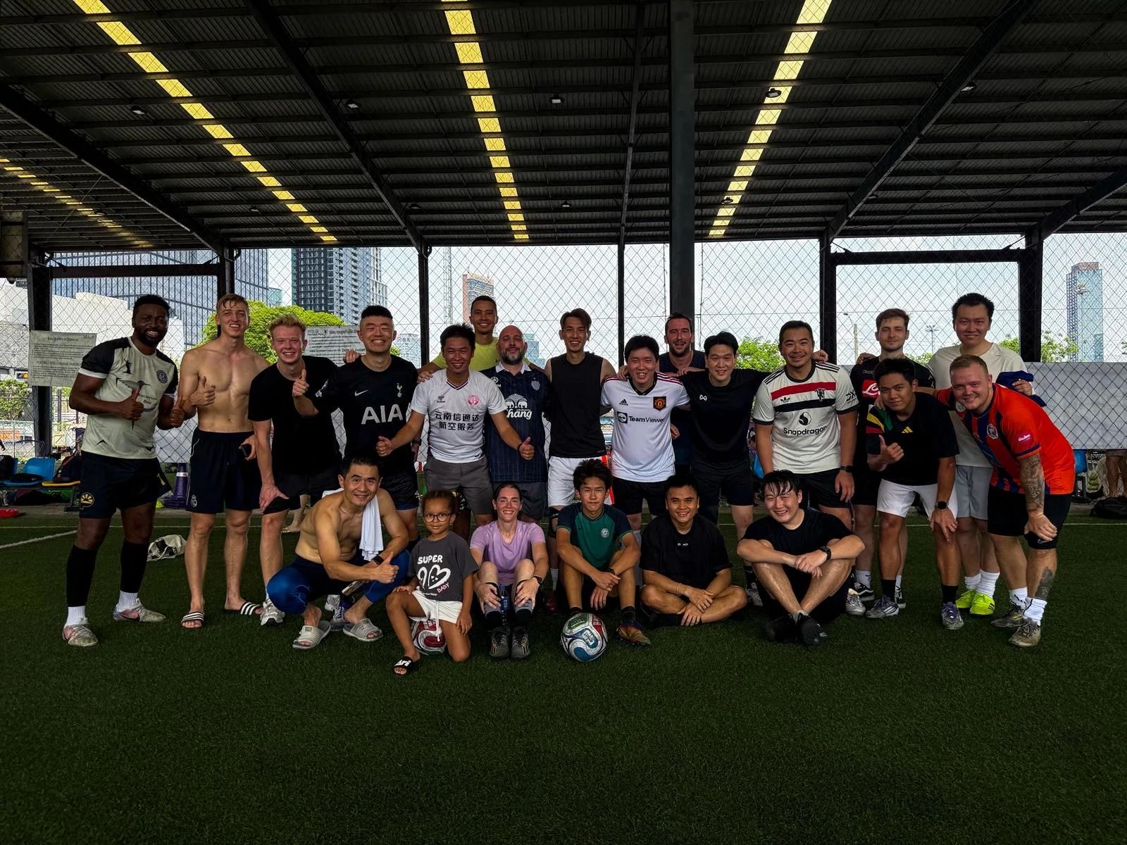 Diverse group of expat football players gathering at POLO Football Park in Bangkok for a pickup football game under floodlights